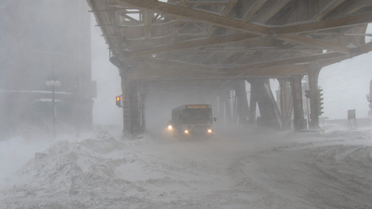 A CTA bus pushing through snow-covered Wacker Drive beneath the Loop's overhead train tracks in downtown Chicago during a whiteout