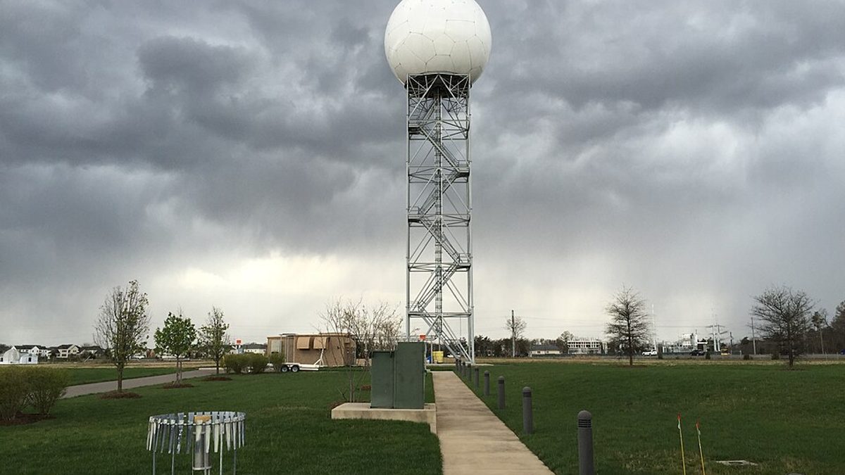 A WSR-88D Doppler radar tower under storm clouds at a National Weather Service forecast office, with an alter-shielded rain gauge and snow boards on the grass below